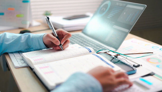 Business professional writing in a planner while working on a laptop, surrounded by reports and charts, representing productivity, time management, business planning, organization, and workflow.
