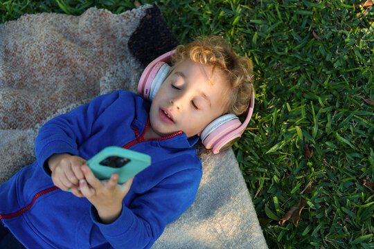 Toddler relaxing outdoors, listening to audio with pink headphones and smartphone on a blanket