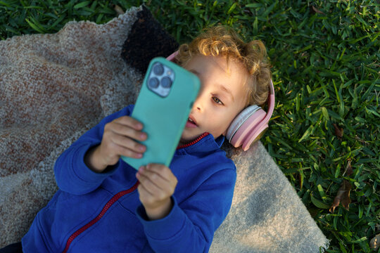 Young child watching media on smartphone and listening with headphones while relaxing on green grass outdoors