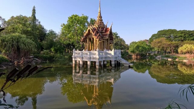 Wide shot of a beautiful Buddhist temple standing on white stilts in a lake, surrounded by trees and mangroves at sunset in a Bangkok park.