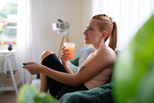 A white woman relaxing on couch with juice and remote control at home