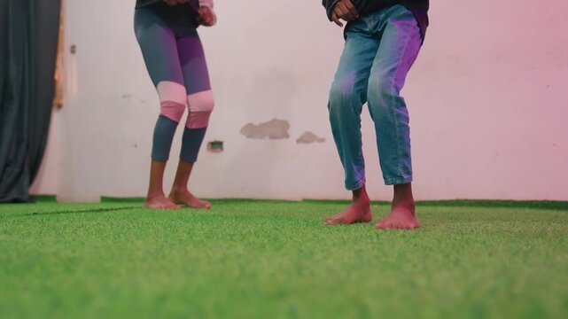 Black mother and child barefoot dancing in lowangle closeup on green turf focus on synchronized steps and footwork with mother as rhythm coach, studio pink lighting and textured grass creating