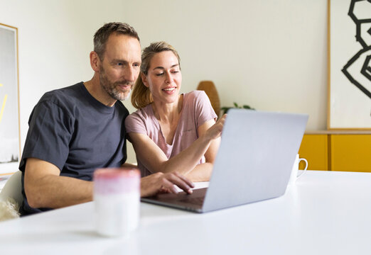 A smiling couple works together on a laptop at a desk indoors