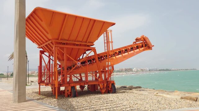 Industrial mobile conveyor and hopper machine beside coastal promenade. Heavy material handling equipment near seaside. Port engineering, bulk cargo transport and industrial machinery infrastructure
