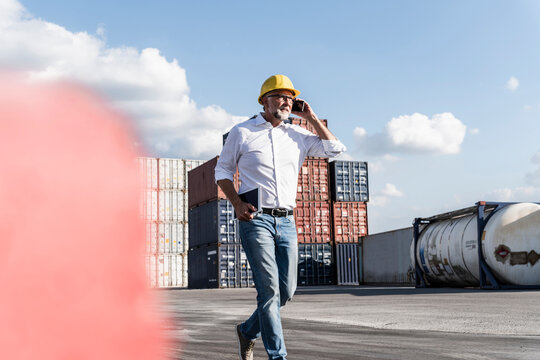 Businessman at cargo harbour, weraing safety helmet, using smartphone and digital tablet