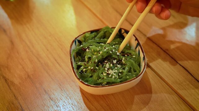 Woman picking chuka seaweed salad with chopsticks at Asian restaurant. Green wakame salad with sesame seeds on wooden table. Japanese cuisine and healthy seafood appetizer close up