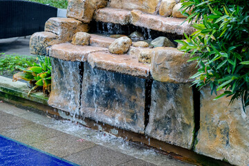Stone waterfall cascades gently over tiered rock formations. Green foliage frames the natural water feature. Water pools softly at the base, reflecting calm surroundings © uniqephoto