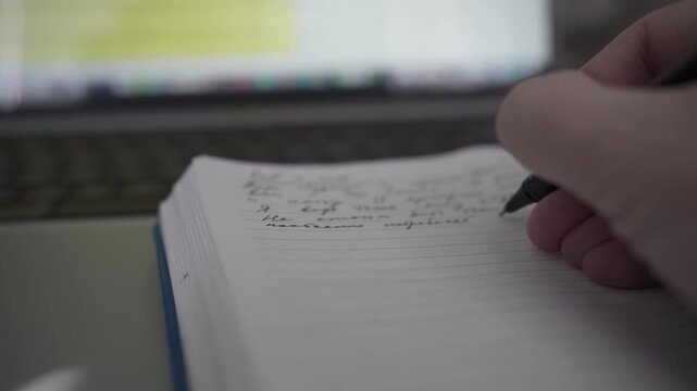 Man taking handwritten notes in notebook while working on laptop at home office desk. Concept of productivity, planning and focused remote work.