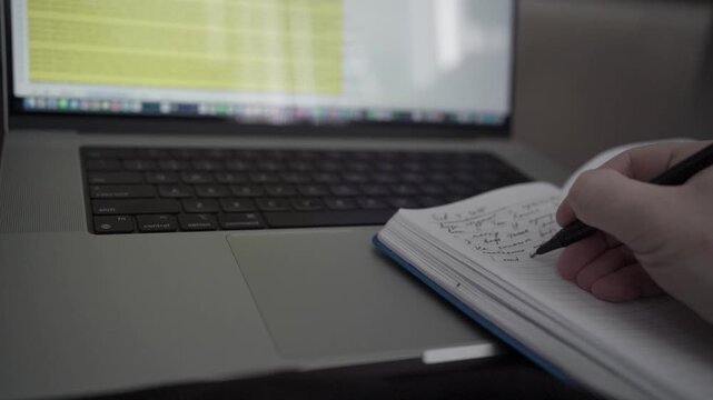 Man taking handwritten notes in notebook while working on laptop at home office desk. Concept of productivity, planning and focused remote work.
