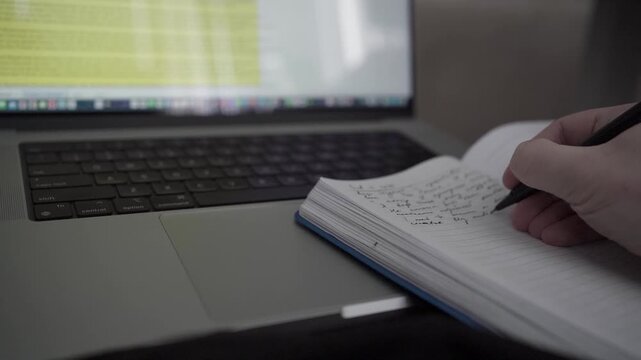 Man taking handwritten notes in notebook while working on laptop at home office desk. Concept of productivity, planning and focused remote work.