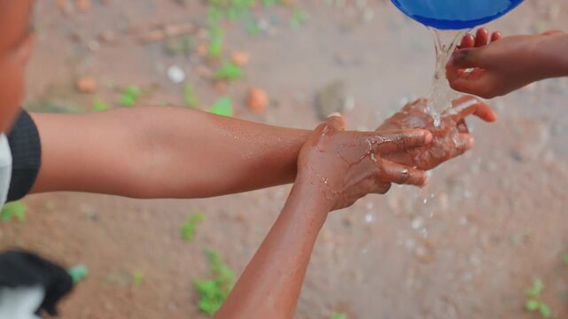 Black mother guiding child hands under pouring water bucket, dusty ground closeup, gentle instruction and hygiene practice, droplets and splash, tender bond between parent and child