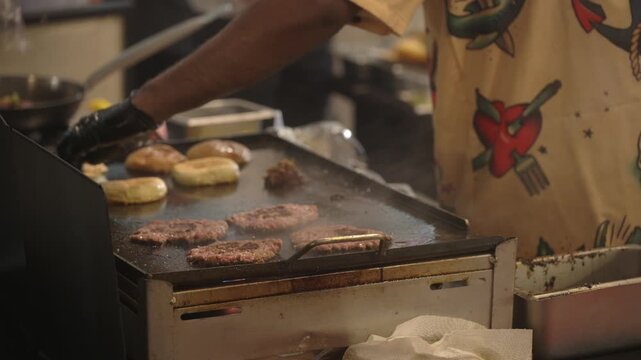 A cook sears beef patties and toasts buns on a hot flat top griddle while preparing fresh burgers inside a busy street food stall