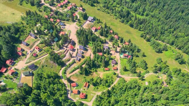 Aerial drone flies over a Balkan mountain village in Serbia, revealing scattered red-roofed houses, winding rural roads, green meadows and dense surrounding forest.