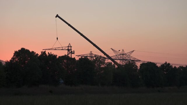 Industrial climbers are working on a power pole with an angle grinder. One arm of the pole is suspended from a truck-mounted crane and is being dismantled as the sun sets.