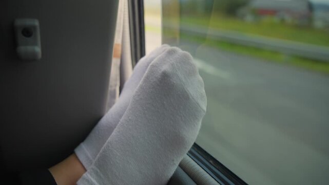 Passenger with feet in white socks resting on a bus or car window, relaxing or sleeping during scenic bus tour journey or commuting between cities or countries. Cars pass on highway in background