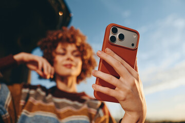 Woman lifestyle outdoors in summer, holding smartphone for selfie, curly hair portrait against sky, casual striped top, relaxed smile, soft bokeh background, daytime street walk © SHOTPRIME STUDIO