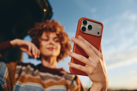 Woman lifestyle outdoors in summer, holding smartphone for selfie, curly hair portrait against sky, casual striped top, relaxed smile, soft bokeh background, daytime street walk