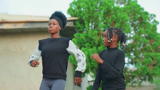 Black mother and child dancing outdoors in neighborhood yard, rhythmic steps and matching black tops, playful coaching and laughter, green tree backdrop, energetic movement and joyful connection