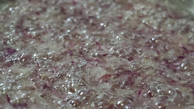 Extreme close up video of finely minced red onions simmering and deep frying in hot transparent oil, showing detailed textures in slow motion movement.

