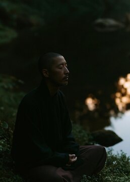 Man sitting on the ground in a meditative pose. he is wearing a black long-sleeved shirt and maroon pants.
