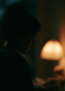 Silhouette of a person sitting at a table with a lamp in front of them. the person appears to be deep in thought, with their head tilted downwards and their eyes closed.