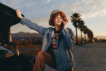 Obraz premium Smiling woman lifestyle outdoors near car at sunset, wearing denim jacket and curly hair, standing on street with warm evening light, relaxed travel pose and sunlit landscape.