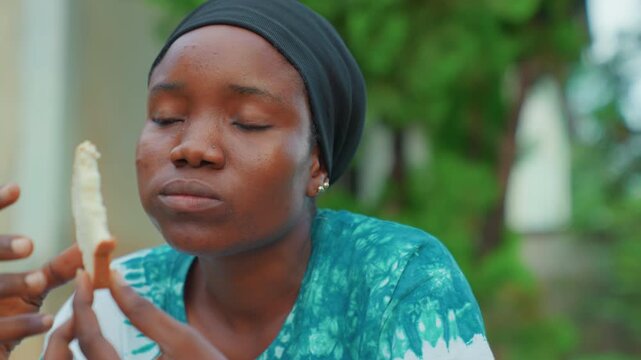 Black mother eating bread alone closeup, headscarf, hands holding torn slice, contemplative expression, outdoor greenery backdrop, natural light, simple everyday nourishment and reflective mood