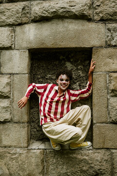 Portrait of smiling Black teenager wearing clown makeup and celebrating Halloween in stone wall niche. Image ideal for seasonal marketing campaigns, vertical