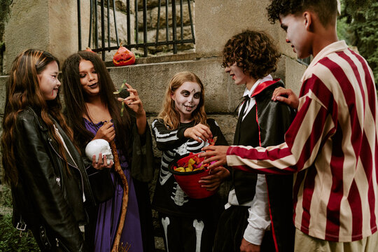 Diverse group of teenagers wearing Halloween costumes sharing candy outdoors. Image suitable for seasonal marketing campaigns, holiday event promotions, and youth lifestyle advertisements