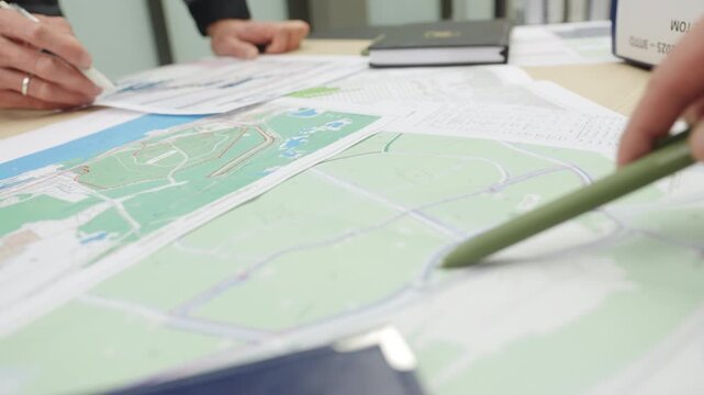 Engineer stands near table covered with colorful regional maps. Specialist wears grey uniform jacket holds white marker pen confidently