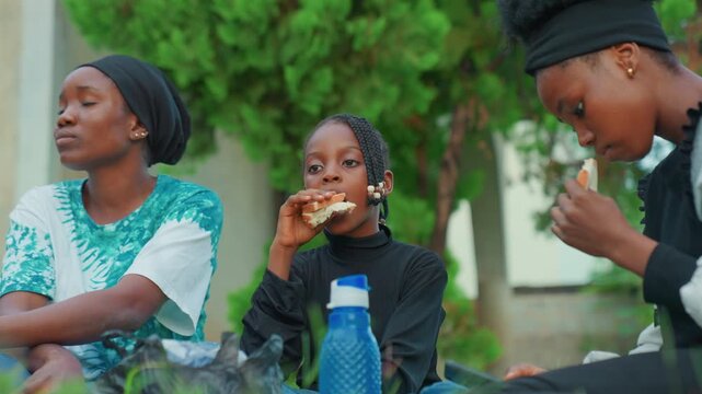 Casual backyard snack african mother child enjoying afternoon together, daughter eating bread while mother leans back in shade, visible water bottle and green foliage, candid family calm with gentle