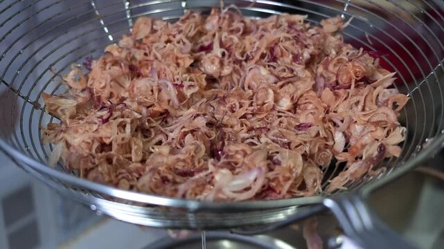 Static slow motion clip of freshly fried golden brown sliced shallots resting on a metal wire strainer, draining oil with additional crispy pieces added from above.
