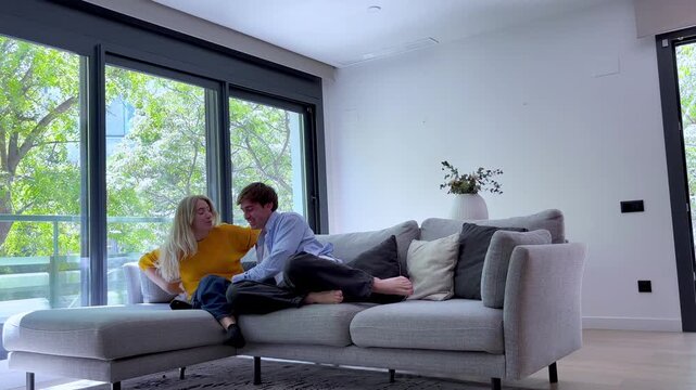 Happy young couple playfully jumping onto the living room sofa, laughing and relaxing together. Cheerful boyfriend and girlfriend having fun, enjoying their quality time at home
