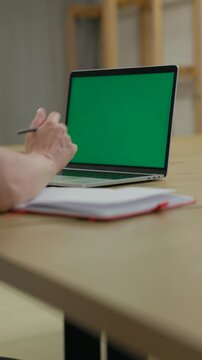 Woman Working On Laptop Computer with Green Mock Up Screen At Home Office Space. Female Freelancer Browsing Green Laptop Screen Writing Information in Notebook. Vertical Video