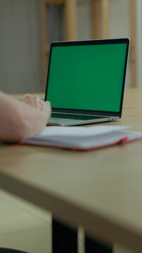 Woman Working On Laptop Computer with Green Mock Up Screen At Home Office Space. Female Freelancer Browsing Green Laptop Screen Writing Information in Notebook. Vertical Video