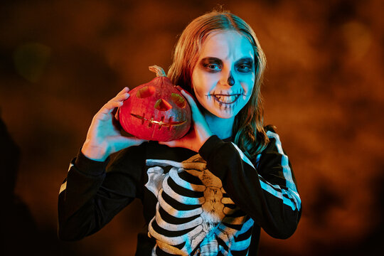 Portrait of Caucasian teenager girl wearing skeleton costume and skull makeup holding carved pumpkin during Halloween celebration, ideal for seasonal marketing campaigns