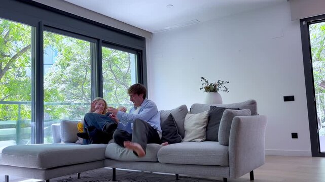 Happy young caucasian couple playfully jumping onto the couch in their bright modern apartment, laughing and talking together while relaxing in the living room with large windows