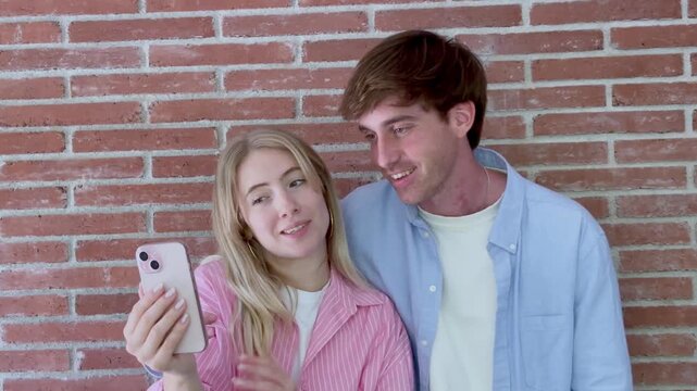 Young happy couple standing against a brick wall, having a fun video call with family or friends using a modern smartphone, smiling and gesturing while communicating online together