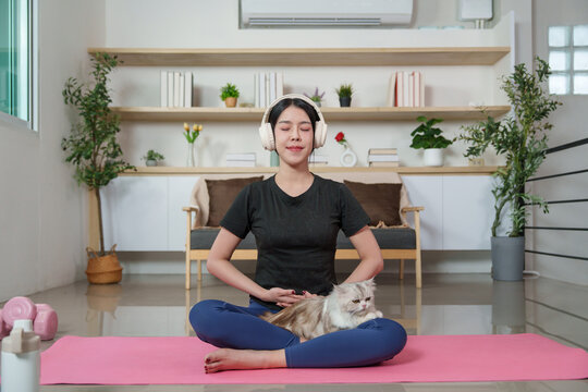 Asian woman meditating with wireless headphones and cat on yoga mat at home