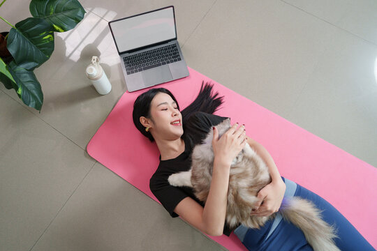 Overhead view of smiling Asian woman hugging cat on yoga mat with laptop