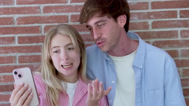 Cheerful young couple standing against a brick wall, happily talking and waving during a video call on a smartphone, connecting with friends or family and sharing a positive moment