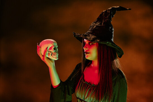 Biracial teenage girl wearing witch costume holding human skull during Halloween celebration. Image suitable for seasonal party invitations and festive marketing campaigns