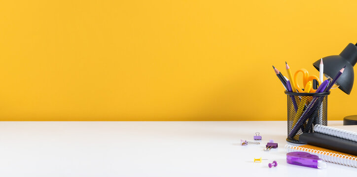 Black Desk Lamp and Yellow Stationery in a Holder on a White Desk Against a Yellow Wall. Minimalist Office Workplace Background with Notebooks and Large Copy Space for Text. Banner Layout.