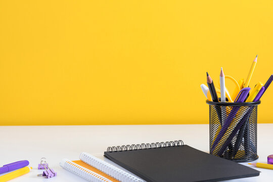 Black Notebook and Yellow Stationery in a Holder on a White Desk Against a Yellow Wall. Minimalist Office Workplace Background with Notebooks, Paper Clips, and Copy Space for Text. Banner Layout.