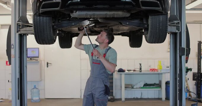 Car mechanic is working under a vehicle on a lift in service. Repairman is using ratchet.