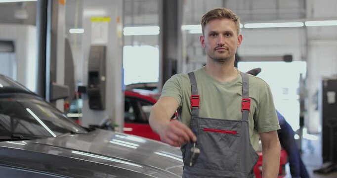Auto mechanic showing car keys at camera. Smiling Caucasian man posing at workplace in repair shop. Automotive industry, auto care, service.