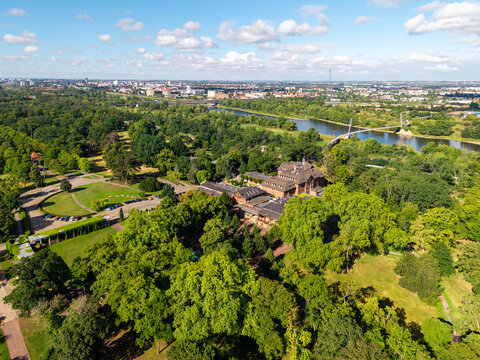Drone view of Herrenkrug pedestrian bridge crossing Elbe River in Magdeburg, surrounded by green parkland and urban scenery. Summer cityscape shows waterfront, trees, architecture and transport links