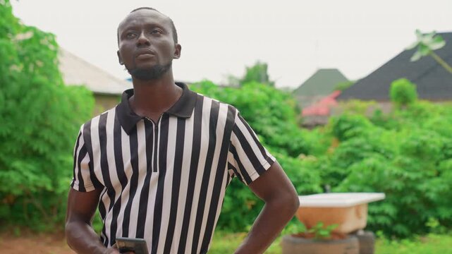 Black man checking phone in backyard. Local referee taking a quiet break in suburban garden, striped polo, casual stance and thoughtful expression, smartphone in hand, soft daylight and lush green