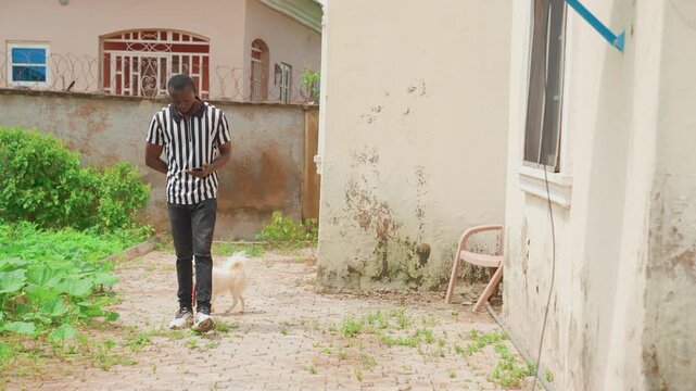Black man checking phone while dog waits in sunlit yard striped shirt, red leash slack at feet, casual stance with pocketed device, subtle companionship and distracted moment in urban courtyard