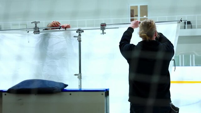 Crew members install diffusion banner in ice rink stadium. Professional technicians prepare photo shooting location stage using metal stands